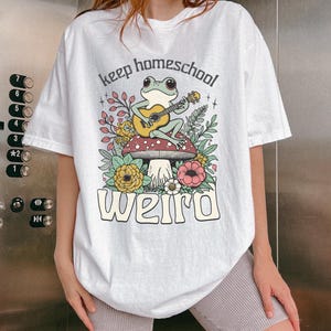May include: White t-shirt with a whimsical graphic featuring a frog playing a guitar while sitting on a mushroom. The text "keep homeschool weird" is printed below the frog illustration. The design includes flowers and foliage.