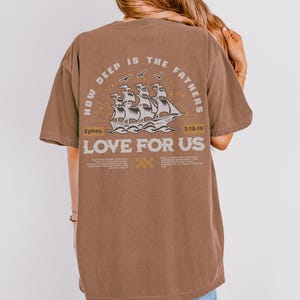 May include: A brown t-shirt with the text "HOW DEEP IS THE FATHERS LOVE FOR US" and a graphic of a ship. The text "Ephes. 3:18-19" is also printed on the shirt. The shirt is made of a soft material.