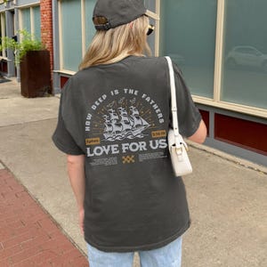 May include: Dark gray t-shirt with the text "HOW DEEP IS THE FATHERS LOVE FOR US" and a ship illustration. The shirt also features the text "Ephes. 3:18-19". The shirt is paired with light blue jeans and a gray baseball cap.
