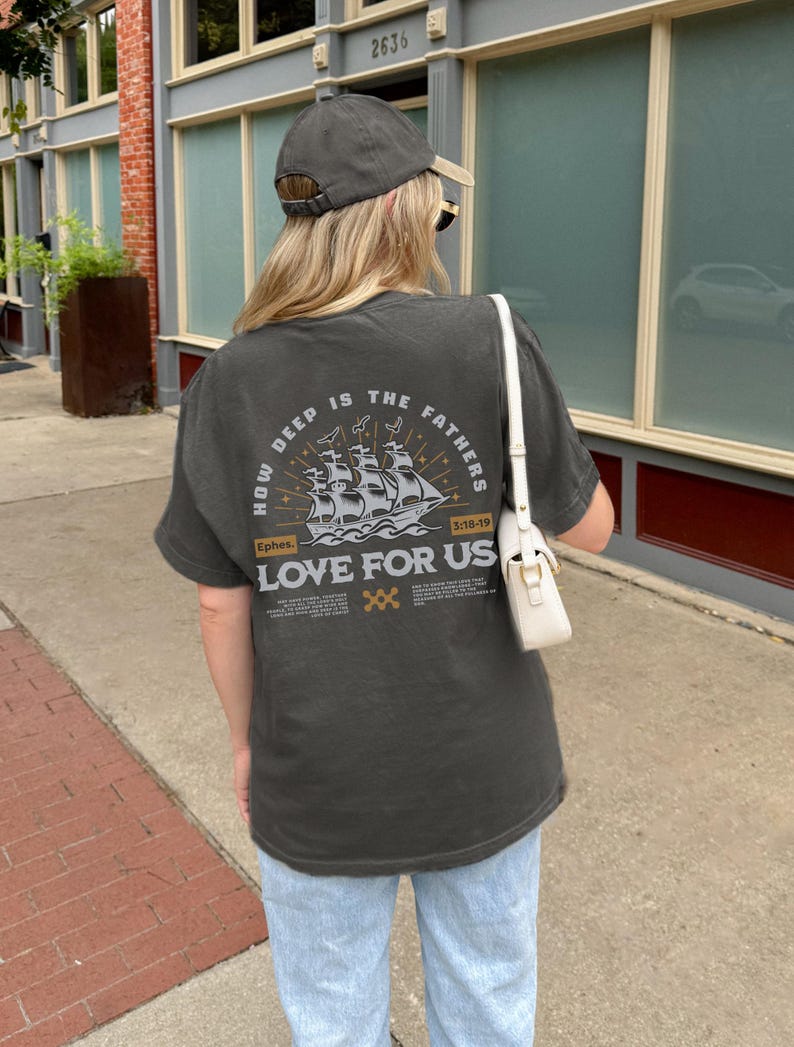 May include: Dark gray t-shirt with the text "HOW DEEP IS THE FATHERS LOVE FOR US" and a ship illustration. The shirt also features the text "Ephes. 3:18-19". The shirt is paired with light blue jeans and a gray baseball cap.