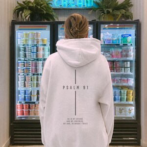May include: A white hooded sweatshirt with the text "PSALM 91" and a vertical line design on the back. Below the text is the phrase "HE IS MY REFUSE AND MY FORTRESS, MY GOD, IN WHOM I TRUST". The person is standing in front of an ice cream display.