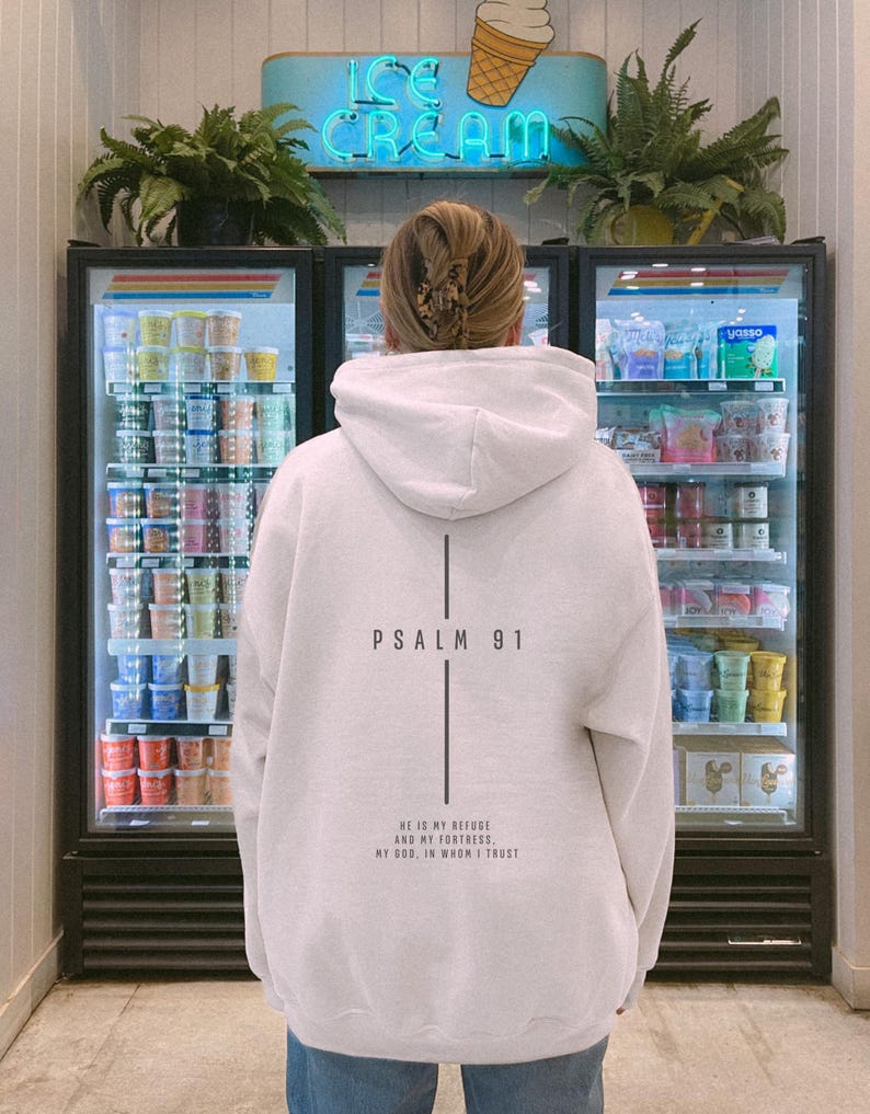 May include: A white hooded sweatshirt with the text "PSALM 91" and a vertical line design on the back. Below the text is the phrase "HE IS MY REFUSE AND MY FORTRESS, MY GOD, IN WHOM I TRUST". The person is standing in front of an ice cream display.