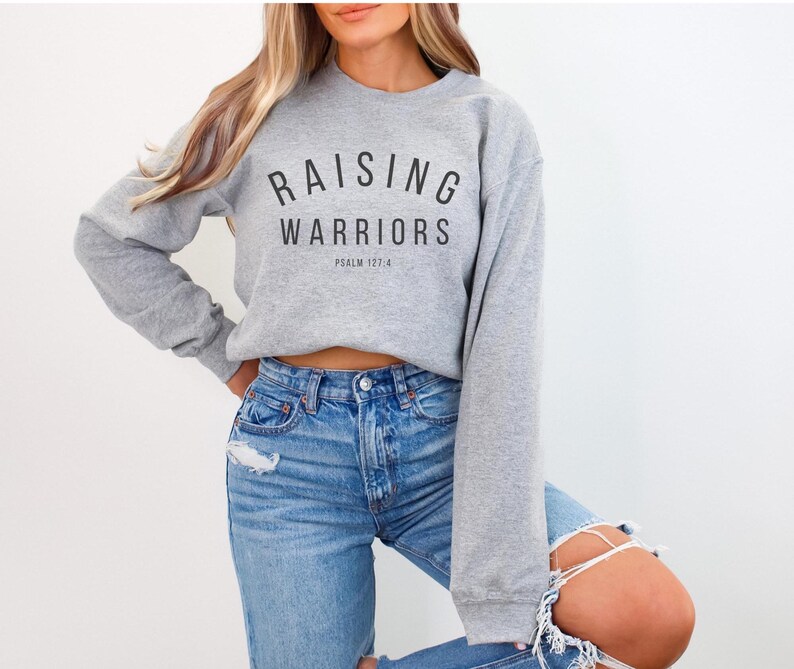May include: A light grey sweatshirt featuring the text "RAISING WARRIORS" in black, with "PSALM 127:4" below. The sweatshirt is worn with light blue denim jeans.