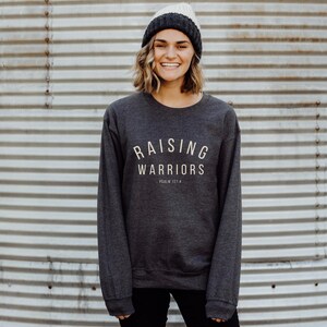 May include: Dark grey crew neck sweatshirt with the text "RAISING WARRIORS" in a light beige colour. The sweatshirt also includes the text "PSALM 127:4". The person in the image is wearing a grey and cream-coloured beanie.