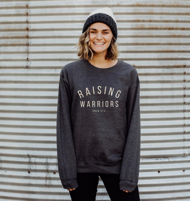 May include: Dark grey crew neck sweatshirt with the text "RAISING WARRIORS" in a light beige colour. The sweatshirt also includes the text "PSALM 127:4". The person in the image is wearing a grey and cream-coloured beanie.