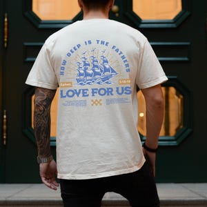 May include: Cream-colored t-shirt with a blue nautical design featuring a ship and the text "HOW DEEP IS THE FATHERS LOVE FOR US". The shirt also includes the text "Ephes. 3:18-19".