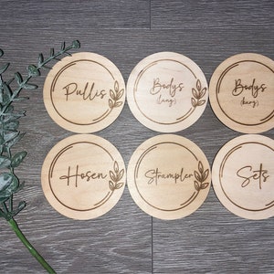 Könnte beinhalten: Sechs runde Holzetiketten mit unterschiedlichem Text und einem Blattdesign. Die Etiketten lauten "Pullis", "Bodys (lang)", "Bodys (kurz)", "Hosen", "Strampler" und "Sets".