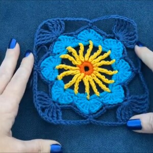 May include: A crocheted square with a blue and yellow flower design. The flower has a yellow center with an orange circle in the middle.