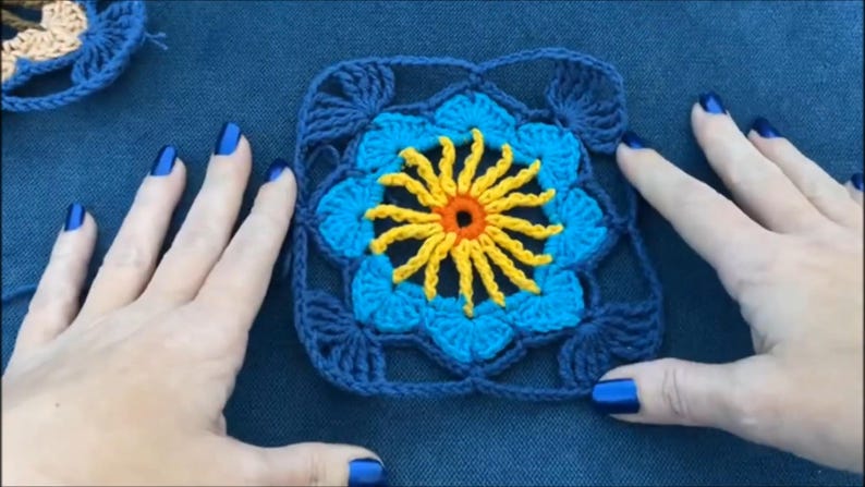 May include: A crocheted square with a blue and yellow flower design. The flower has a yellow center with an orange circle in the middle.