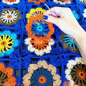 May include: A close-up of a crocheted blanket with a colorful floral pattern. The blanket is made of blue, orange, yellow, white, and brown yarn. A hand is holding a crochet hook and working on the blanket.