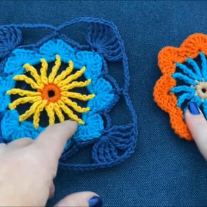 May include: Two crocheted flower motifs, one blue and yellow and one orange and blue, with a yellow center and a blue center, respectively. The motifs are on a blue background.