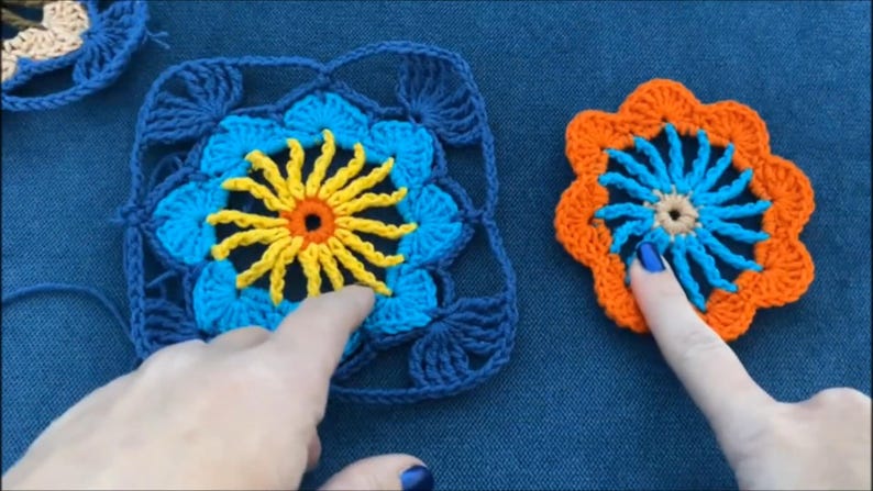 May include: Two crocheted flower motifs, one blue and yellow and one orange and blue, with a yellow center and a blue center, respectively. The motifs are on a blue background.