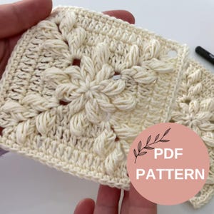 May include: A close-up of a crocheted square with a floral design in a cream colour. The square is made with a textured stitch and has a hole in the centre. The text "PDF PATTERN" is visible in a circle with a floral design.