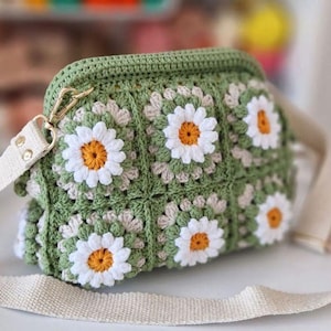 Könnte beinhalten: Eine grüne gehäkelte Tasche mit einem weißen Gänseblümchenmuster und einem beigefarbenen Riemen. Die Tasche hat einen goldenen Verschluss.