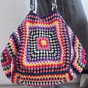 May include: A colorful crocheted tote bag with a large, intricate granny square pattern in shades of pink, purple, yellow, and green. The bag has a dark brown crocheted border and two brown leather handles.