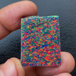 Könnte beinhalten: Ein rechteckiger Edelstein mit einem lebendigen Mosaik aus Farben, darunter Rot, Orange, Gelb, Grün, Blau und Lila. Der Stein wird in einer Hand gehalten, wodurch seine polierte Oberfläche und das komplizierte Muster zur Geltung kommen. Der Edelstein scheint ein Opal zu sein.