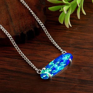 May include: A silver chain necklace with a rectangular pendant. The pendant features a vibrant blue and green opal stone. The necklace is displayed on a dark wooden surface, with a sprig of green leaves in the background.