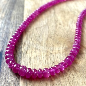 May include: A strand of small, round, vibrant fuchsia-pink gemstone beads arranged in a curved line. Each bead features a smooth, polished surface with subtle variations in color and translucency, creating a continuous, uniform string of beads.