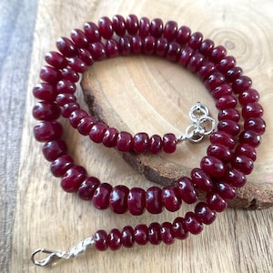 May include: A necklace composed of numerous small, smooth, deep red circular beads strung together. The strand features a polished silver-toned metal lobster claw clasp and a circular jump ring connector at the ends, securing the circular arrangement.