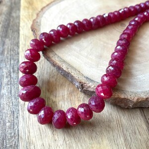 May include: A necklace featuring faceted, deep red gemstones. The oval-shaped beads are strung together, forming a continuous loop. The necklace is presented on a wooden surface, highlighting the rich colour and texture of the stones.