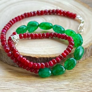 Puede incluir: Un collar con hebras alternas de pequeñas cuentas rojas facetadas y cuentas verdes ovaladas más grandes. El collar se asegura con un cierre plateado. La joya descansa sobre una superficie de madera, mostrando los colores vibrantes y el diseño.
