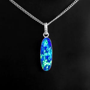 May include: A silver necklace with an oval pendant. The pendant features a vibrant blue and green opal stone. The chain is a delicate silver link. The background is solid black, highlighting the jewelry.