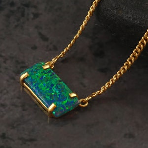 May include: A gold necklace featuring a rectangular pendant with a vibrant, iridescent opal. The opal displays shades of green and blue. The pendant is encased in a gold setting and hangs from a delicate gold chain. Jewelry.