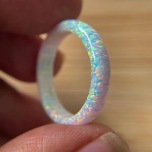May include: A circular ring featuring a smooth, polished surface with an iridescent opal-like finish. The band displays a vibrant spectrum of light blue, pink, and yellow flecks that shift in color across the rounded, continuous surface.