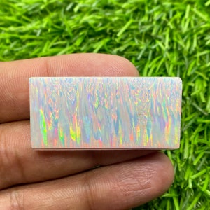 May include: A white opal cabochon with iridescent rainbow colours. The opal is rectangular and has a smooth, polished surface.