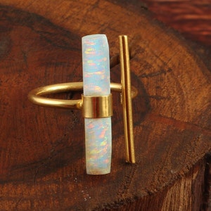 Könnte beinhalten: Ein goldfarbener Ring mit einem vertikalen, rechteckigen Opalstein mit irisierenden Farbtönen. Ein vertikaler Goldstab erstreckt sich vom Ringband und erzeugt ein modernes, geometrisches Design. Der Ring liegt auf einer dunklen Holzoberfläche.