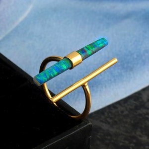 May include: A gold-colored ring featuring a vibrant, rectangular opal stone with blue and green hues. The opal is set horizontally, held by a gold band and a vertical gold bar, creating a modern, geometric design.