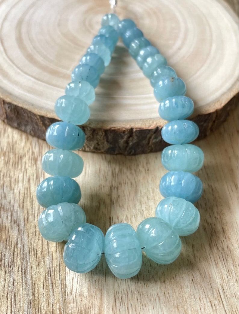 May include: A strand of light blue aquamarine beads, each bead is carved with a textured design. The beads are strung on a silver thread.