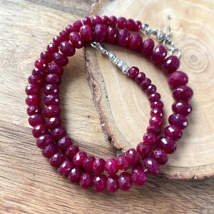 May include: A necklace composed of small, faceted, deep red ruby beads arranged in a circular pattern. The strand features a silver-toned metal lobster claw clasp and a short extender chain with small silver-toned beads at the end of the closure.