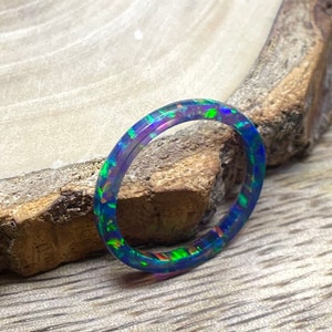Könnte beinhalten: Ein dünner, blauer Ring mit schillernden grünen, violetten und rosafarbenen Flecken. Der Ring ist aus Harz gefertigt und ein einzigartiges Schmuckstück.