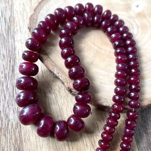 May include: A strand of deep red, polished, round beads arranged in a curved shape. The beads vary slightly in size, with larger beads concentrated on one side of the strand, transitioning to smaller, uniform beads along the rest of the length.