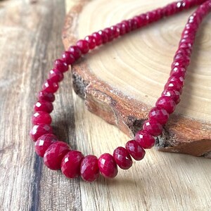 May include: A necklace made of deep red faceted gemstone beads. The beads are strung on a clear thread and the necklace is lying on a wooden surface.