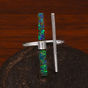 Op de afbeelding: Een zilveren ring met een verticale rechthoekige opaalsteen met blauwe, groene en zwarte tinten. Een parallelle zilveren staaf loopt van de ringband. De ring ligt op een donker, getextureerd oppervlak.