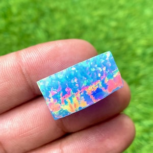 Puede incluir: Una piedra preciosa de ópalo rectangular e iridiscente con una vibrante exhibición de colores. El ópalo presenta una base azul con destellos de rosa, amarillo y naranja. La piedra preciosa se sostiene en una mano sobre un fondo verde borroso.