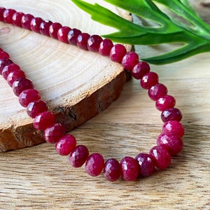 Puede incluir: Una hebra de cuentas de piedras preciosas facetadas de color rojo intenso. Las cuentas son redondas y tienen un acabado pulido. Las cuentas están enhebradas en un hilo transparente y se muestran sobre una superficie de madera.