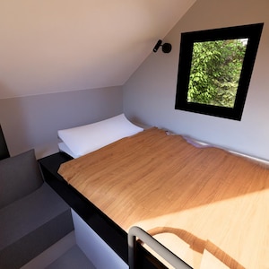 以下が含まれることがあります： ベッドと作り付けのソファを備えた、小さくモダンな寝室のインテリア。ベッドには木製のヘッドボードと白い枕があります。黒いフレームの窓からは緑の葉が見えます。部屋はニュートラルな色調で塗装されています。