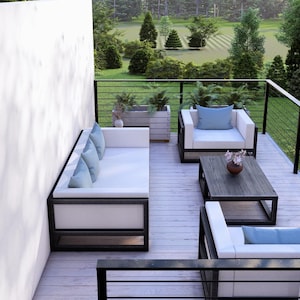 May include: Modern outdoor patio furniture set. The set features a white sofa, two armchairs, and a coffee table with black frames. Light blue cushions and pillows provide colour. The patio has a wooden deck and a black railing, with a garden view.
