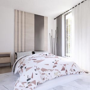 Op de afbeelding: Een modern slaapkamerinterieur met een bed met een patroon dekbed in wit, bruin en grijstinten. De kamer heeft een houten hoofdbord, een nachtkastje en een groot raam met witte gordijnen. De muren zijn wit geschilderd.