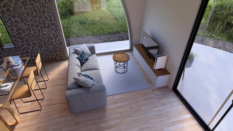 May include: An interior shot of a modern living room with a gray sofa, a round coffee table, and a wall-mounted TV. The room has large windows overlooking a green outdoor area. A dining table with chairs is also visible.