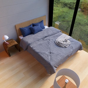 May include: A bedroom with a wooden bed frame, blue pillows, and a gray comforter. A small wooden side table holds a white lamp. The room has large windows overlooking a green landscape and wooden floors.
