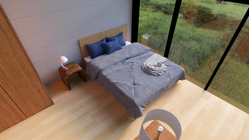 May include: A bedroom with a wooden bed frame, blue pillows, and a gray comforter. A small wooden side table holds a white lamp. The room has large windows overlooking a green landscape and wooden floors.