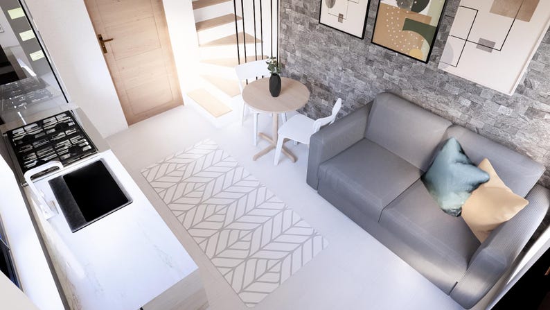 May include: Interior view of a compact living space with a kitchen area. A grey sofa with cushions is against a stone wall. A small round table and two white chairs are near a patterned rug. A hob and sink are visible.