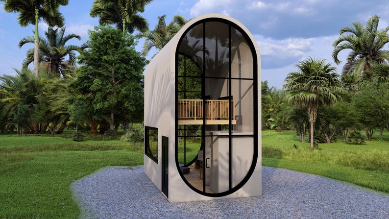 May include: A modern, two-story tiny house with a unique oval window design. The structure features a light-colored exterior, large glass windows, and a small balcony. The house is surrounded by lush green trees and grass, with a gravel path leading up to it.
