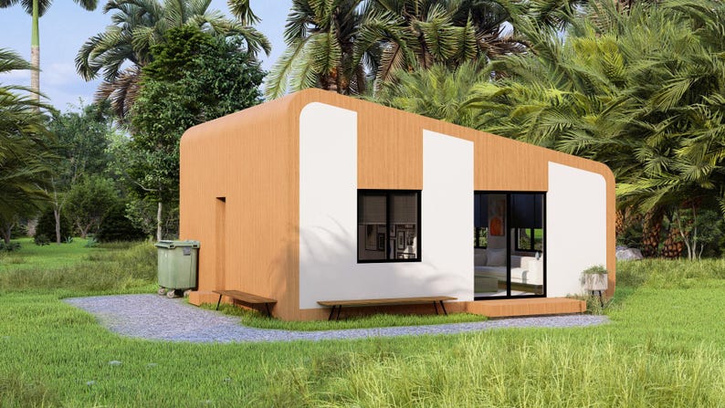 Puede incluir: Una caba&ntilde;a moderna de una sola planta con un exterior marr&oacute;n claro y paneles blancos. La caba&ntilde;a tiene ventanas grandes, un peque&ntilde;o banco y un cubo de basura verde. Est&aacute; rodeada de exuberante hierba verde y follaje tropical.