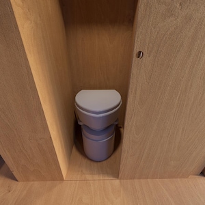 Pu&ograve; includere: Un WC a compostaggio grigio chiaro &egrave; inserito in un involucro di legno. Il WC ha un sedile arrotondato e una base cilindrica. Le pareti e la porta circostanti sono in legno chiaro, creando un'estetica minimalista.