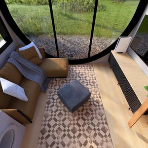 May include: Interior view of a modern living space with large windows overlooking a green landscape. The room features a brown sectional sofa with white pillows, a patterned rug, and a wooden ladder. A small gray ottoman sits on the rug.